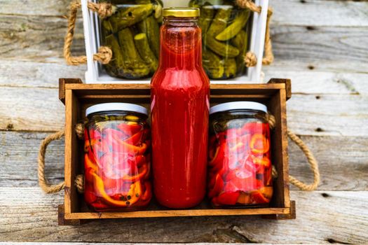 Wooden crate with bottles with tomatoes sauce and glass jars with pickled red bell peppers isolated in a rustic composition. Jars with variety of pickled vegetables preserved food concept.