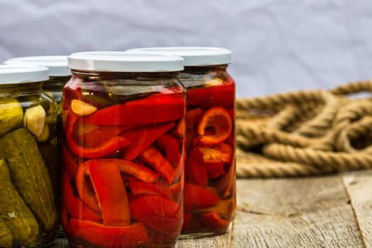 Glass jars with pickled red bell peppers and pickled cucumbers (pickles) isolated. Jars with variety of pickled vegetables. Preserved food concept in a rustic composition.