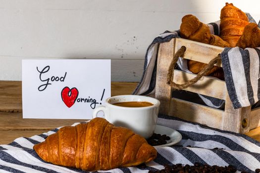 Coffee cup and buttered fresh French croissant on wooden crate. Food and breakfast concept. Morning message “good morning” on white board