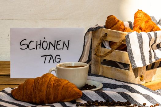 Coffee cup and buttered fresh French croissant on wooden crate. Food and breakfast concept. Morning message in German meaning “have a nice day” on white board