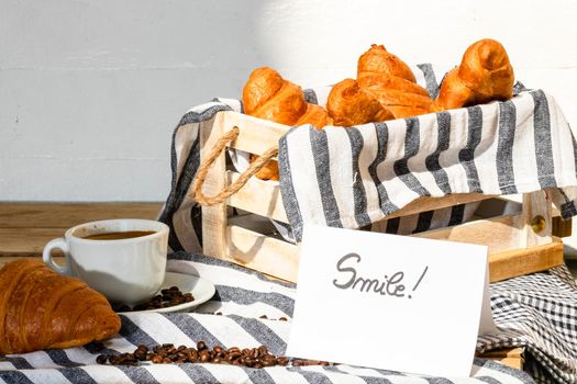 Coffee cup and buttered fresh French croissant on wooden crate. Food and breakfast concept. Morning message “smile” on white board