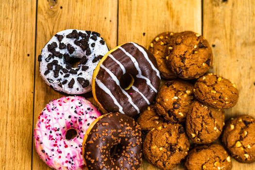 Colorful donuts and biscuits on wooden table. Sweet icing sugar food with glazed sprinkles, doughnut with chocolate frosting. Top view with copy space