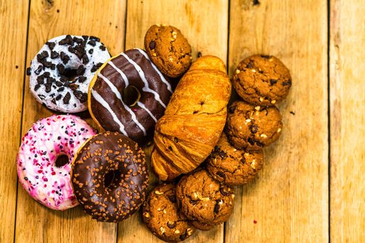 Colorful donuts, puff pastry and biscuits on wooden table. Sweet icing sugar food with glazed sprinkles, doughnut with chocolate frosting. Top view with copy space