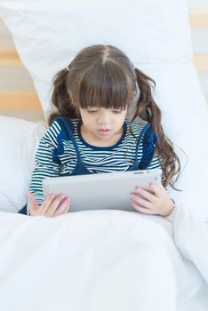 Cute asian little girl enjoy watching cartoon on smart tablet while sitting on bed in kid bedroom at home
