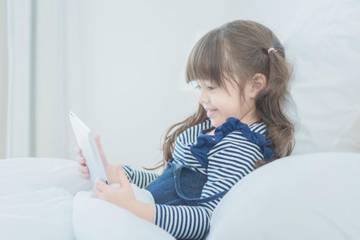 Cute little girl enjoy watching cartoon on smart tablet with cute doll while sitting on bed in kid's bedroom at home