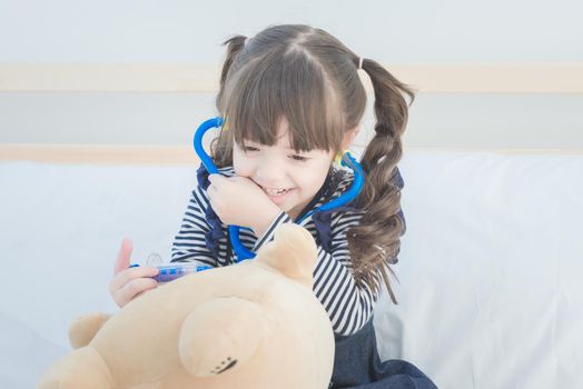 Cute little girl enjoy playing doctor with doctor toy set and cute doll while sitting on bed in kid's bedroom at home.Elegant design for occupation job,kid playing and healthcare concept