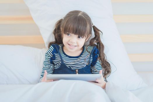 Cute kid girl smiling while using digital smart tablet on bed in kid's bedroom at home.Photo design for Happy Family and kids concept.