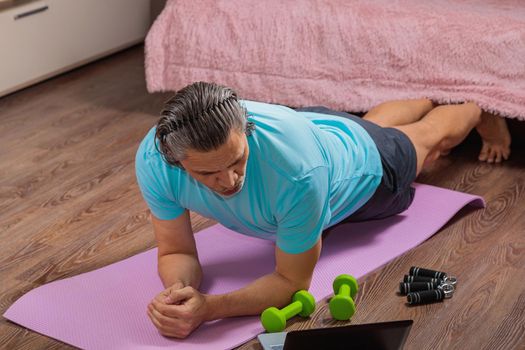 50-year-old man performs exercises while lying on the rug at home, looking at the computer. During a pandemic, a person trains in an apartment via the Internet.