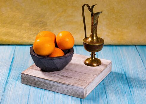 A group of oranges in a concrete geodesic bowl and a etched brass urn on a cyan base with light yellow-orange backdrop.