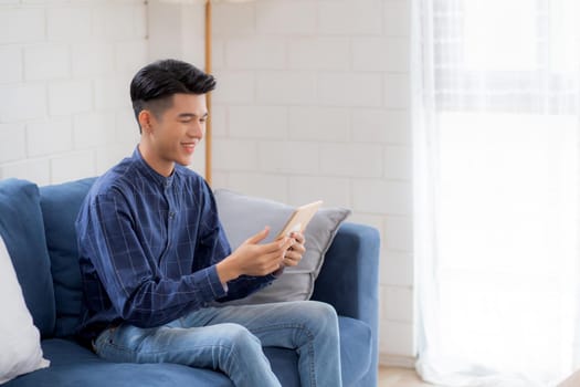 Young attractive asian man resting using browsing tablet computer on sofa at home, happy male sitting on couch relax reading digital gadget at house, communication and lifestyle concept.