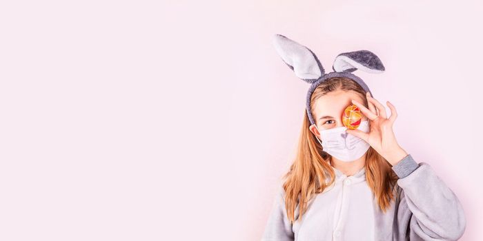 Girl in rabbit bunny ears on head and protective mask with colored eggs on pink background. Cheerful smiling happy child. Covid Easter holiday banner