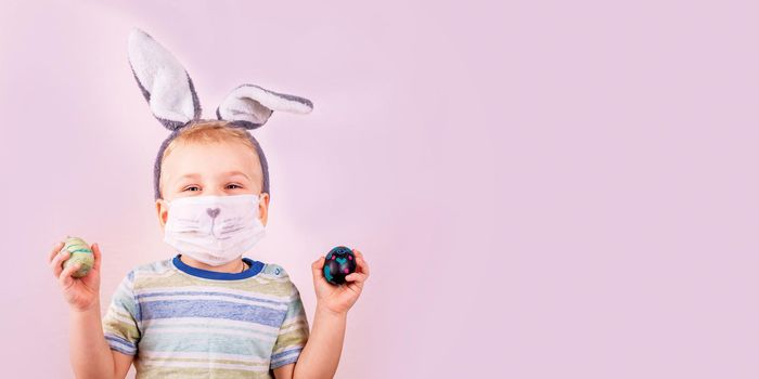 Cute baby boy in rabbit bunny ears on head and protective mask with colored eggs on pink background. Cheerful smiling happy child. Covid Easter holiday banner