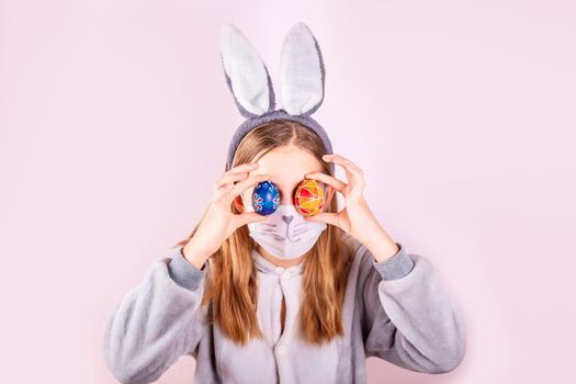 Girl in rabbit bunny ears on head and protective mask with colored eggs on pink background. Cheerful smiling happy child. Covid Easter holiday