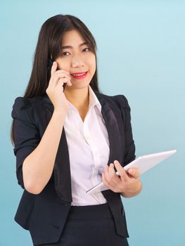 Young Asian women in suit standing using her phone and holding digital tablet against blue background