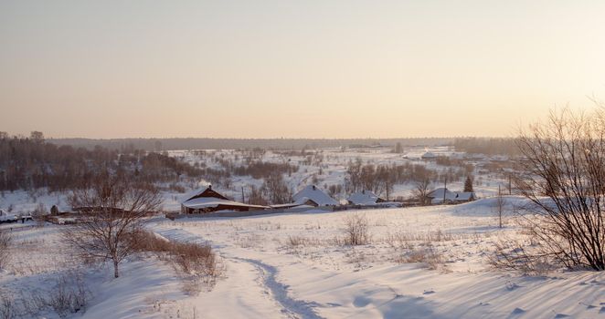 A snow-covered village with houses and a vered road leading to it. There is a lot of snow in the fields and the roofs of the houses barely stick out of the snow