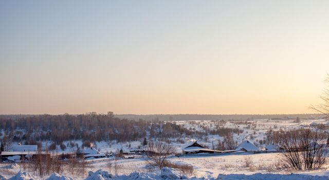 A snow-covered village with houses and a vered road leading to it. There is a lot of snow in the fields and the roofs of the houses barely stick out of the snow