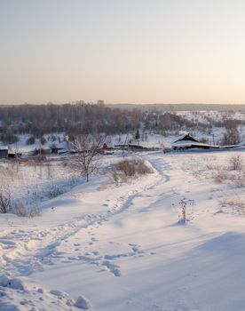 A snow-covered village with houses and a vered road leading to it. There is a lot of snow in the fields and the roofs of the houses barely stick out of the snow