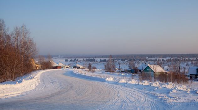 A snow-covered village with houses and a vered road leading to it. There is a lot of snow in the fields and the roofs of the houses barely stick out of the snow