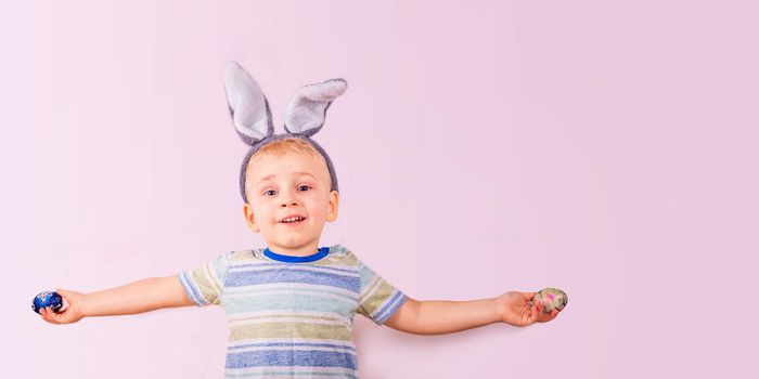 Cute baby boy in rabbit bunny ears on head and with colored eggs on pink background. Cheerful smiling happy child. Easter holiday banner