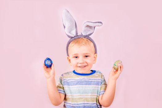 Cute baby boy in rabbit bunny ears on head and with colored eggs on pink background. Cheerful smiling happy child. Easter holiday