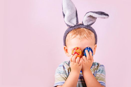 Cute baby boy in rabbit bunny ears on head closing his eyes with colored eggs on pink background. Cheerful smiling happy child. Easter holiday