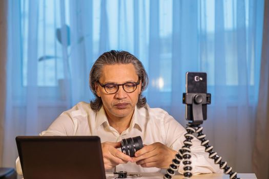 A 50-year-old blogger with glasses is recording an online seminar on his smartphone for his subscribers. With the help of modern devices, a computer and a smartphone, the master class is broadcast live.