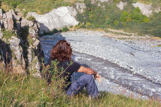 A male traveler with long hair sits on the side of a mountain and looks out at a stormy river.