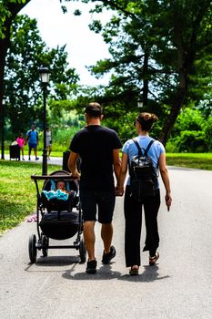 Back view of couple parents holding hands with baby in stroller walking, relaxing and having fun on the alleys of park and gardens of the domain from Mogosoaia in Bucharest, Romania, 2020.