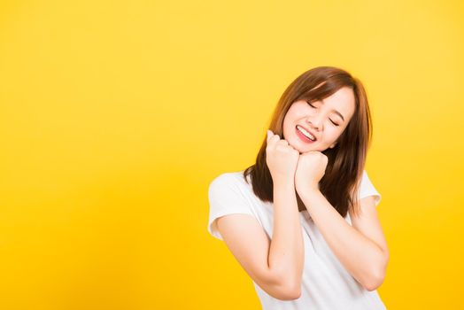 Asian happy portrait beautiful cute young woman teen stand wear t-shirt happy expression fist pressed together under chin and closed eyes isolated, studio shot on yellow background with copy space