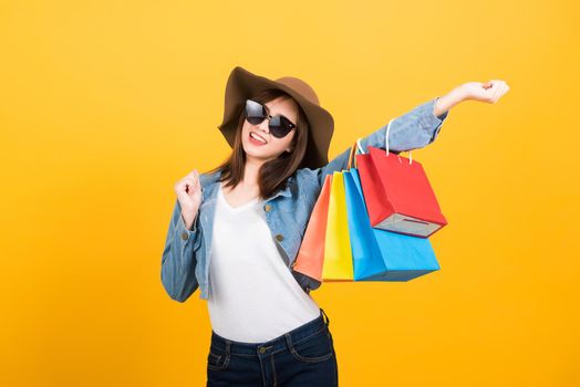 Asian happy portrait beautiful cute young woman teen smiling standing with sunglasses excited holding shopping bags multi color looking camera isolated, studio shot yellow background with copy space