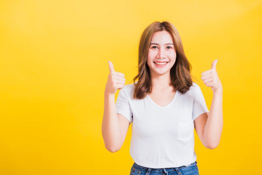 Portrait Asian Thai beautiful happy young woman smiling wear white t-shirt standing successful woman giving two thumbs up gesture sign in studio shot, isolated on yellow background with copy space
