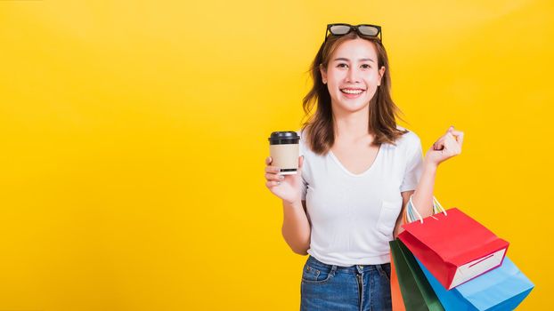 Portrait Asian Thai beautiful happy young woman smiling hold shopping bags multi-color and take away coffee cup her looking to camera, studio shot isolated on yellow background, with copy space