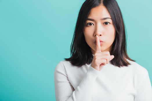 Young beautiful Asian woman holding index finger on her mouth lips, Portrait female hush silence, studio shot isolated on a blue background, Gesture of shhh