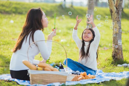 Happy Asian mother and little girl daughter child having fun and enjoying outdoor together sitting on the grass blowing soap bubbles during a picnic in the garden park on a sunny day family time