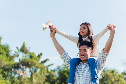 Happy Asian young family father and carrying an excited girl on shoulders having fun and enjoying outdoor lifestyle together playing aircraft toy on sunny summer day, Father's day concept