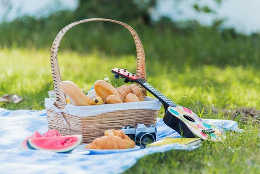 Picnic wattled basket with bread food and fruit, Ukulele, a retro camera on blue cloth in green grass garden with copy space at sunny summertime