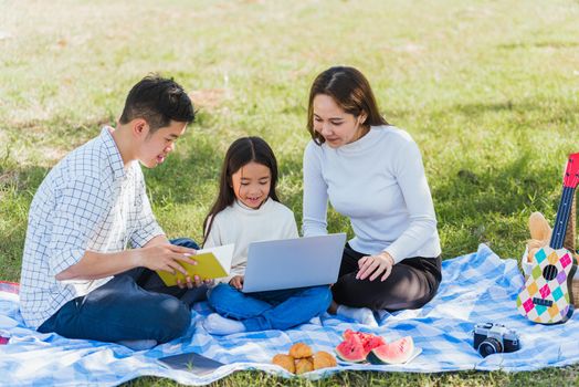 Happy Asian lifestyle young family father, mother and little girl having fun outdoor sitting on picnic blanket using laptop computer technologies while relaxing to open song at garden park in summer