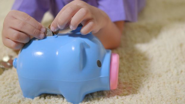 Happy Asian kid boy preschool putting pin money coins into blue faced piglet slot. Little child putting coin into piggy bank for saving with pile of coins at home, Investment education
