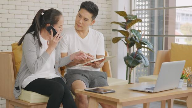 Happy Asian family couple husband and wife working confirming booking having talking call phone conversation and making writing notes of information together from home in living room. small business