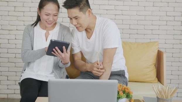 Happy Asian family couple husband and wife work and using digital tablet for discuss. Cheerful loving couple smile shopping something online and watching social media together from home in living room