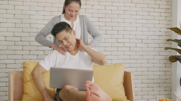 Happy Asian family couple husband and wife laughing sitting on sofa using laptop computer working from home. Business man sitting on sofa home working on laptop her woman is giving her shoulder massage