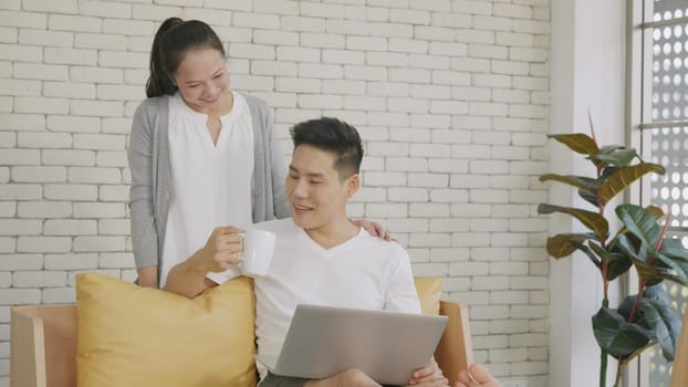 Happy Asian beautiful family couple husband and wife laughing sitting on sofa in the living room working with laptop computer at home. Woman brings coffee to the man during work