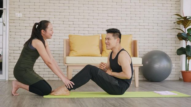 Happy Asian family couple husband and wife healthy trainer workout home in the living room. Fit man lifestyle doing sit up fitness exercise at home on floor with girlfriend support helping boyfriend