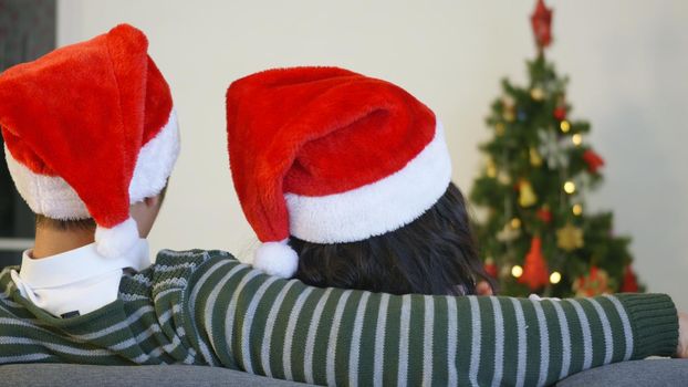 Happy Asian beautiful young family couple husband and wife sitting on sofa wearing a santa hat in front of fireplace at warm home Christmas winter hugging living room, back view,