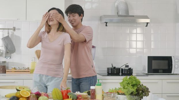Happy Asian beautiful family couple husband and wife cooking salad in kitchen together at home. The man close eyes woman for surprise salad food he made. healthy food concept