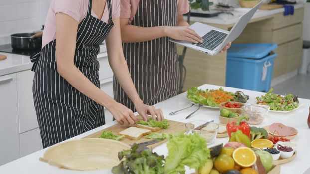 Happy Asian beautiful young family couple husband and wife cooking food vegetable salad in kitchen together at home. The man and woman according online cooking class to recipe on laptop computer