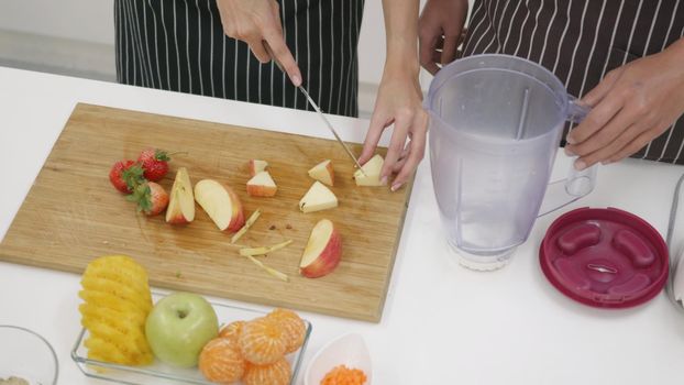 Happy Asian beautiful young family couple husband and wife making fresh smoothie in kitchen together at home. The man and woman slicing fresh apple fruits and veggies. Healthy lifestyle concept