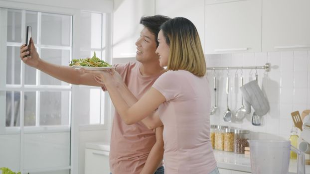 Happy Asian beautiful young family couple husband and wife enjoying smile and laugh, woman and man using smartphone live streaming while salad food spending time together in kitchen at home