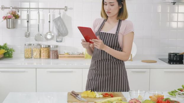 Happy Asian beautiful young woman in kitchen following recipe on digital tablet computer. Female cooking salad online video clip on website via tablet content on internet technology. modern food