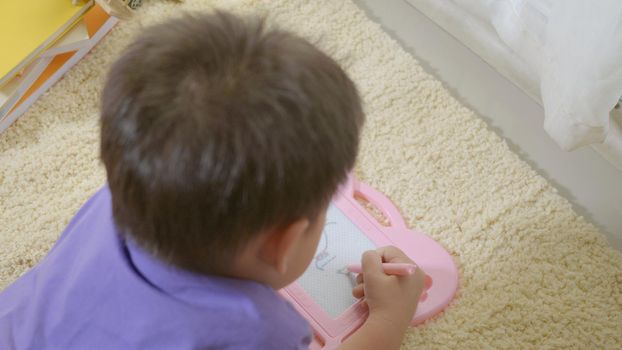 Happy Asian klid little boy preschool writing at the magnetic drawing board at home on carpet. Funny child playing magnetic drawing board. Education Learning to draw concept. Back view